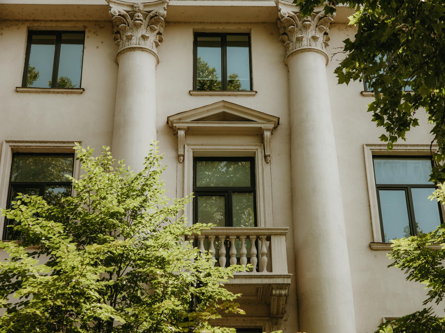 Elewacja neoklasycznej kamienicy z kolumnami otoczona letnimi drzewami